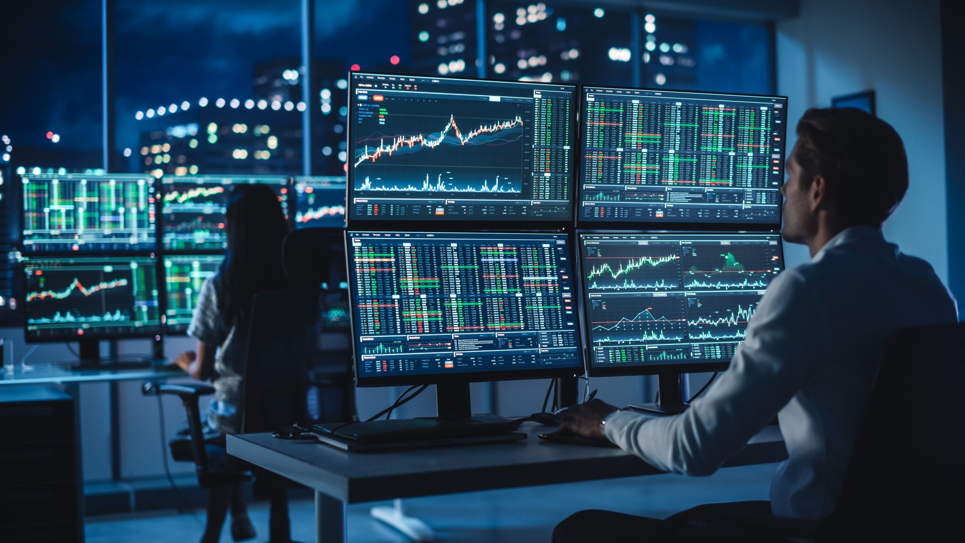 Financial Analyst Working on a Computer with Multi-Monitor Workstation with Real-Time Stocks, Commodities and Foreign Exchange Charts. Businessman Works in Investment Bank City Office at Night.